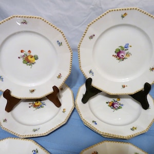 Puede incluir: Un juego de ocho platos de cena de porcelana blanca con ribete dorado y diseños florales. Los platos están apilados uno encima del otro y se muestran sobre una superficie azul.