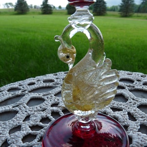 May include: A red and clear glass swan-shaped wine goblet with gold accents. The goblet is sitting on a white crocheted doily.