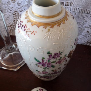 May include: A white porcelain vase with a gold rim and floral designs in shades of purple, pink, and green. The vase has a delicate, hand-painted look.