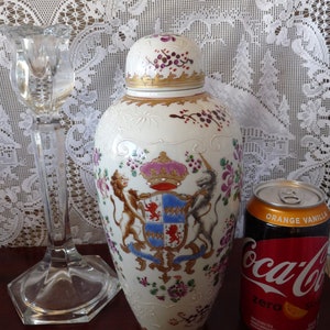 May include: A white porcelain vase with a gold trim and a colorful floral design. The vase features a coat of arms with a lion and a unicorn. The vase is sitting on a table with a white lace tablecloth.