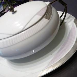 May include: White porcelain soup tureen with a lid and a silver knob. The tureen has a silver trim and a pink and silver design. The tureen is sitting on a matching plate.