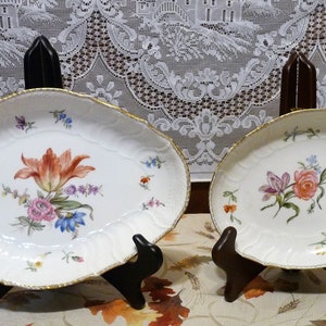 May include: Two white oval porcelain serving dishes with gold trim and floral designs. The larger dish features a large orange tulip with pink and blue flowers. The smaller dish features a pink rose with white and purple flowers.
