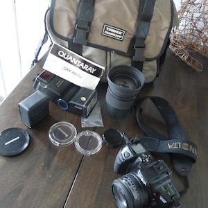 May include: A collection of photography equipment, including a camera, lenses, a flash, and a carrying bag. The camera is black with a lens attached. The bag is tan with black straps and the text "Tundra Chameleon".