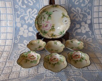 May include: A set of seven vintage porcelain bowls with floral designs. The bowls are in a cream color with pink and green flowers. The largest bowl is on a stand and the smaller bowls are arranged around it.