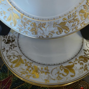 May include: Two white ceramic plates with gold floral designs. The plates are stacked on top of each other and are on a black stand.