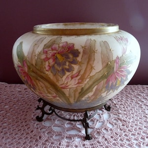 May include: A large, white ceramic bowl with a gold rim and floral design. The bowl is decorated with pink, purple, and green cactus flowers. The bowl is sitting on a metal stand with three legs.