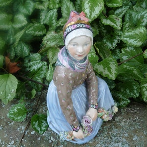 Puede incluir: Una figurita de porcelana de una niña con un vestido azul y un sombrero blanco y rosa, sosteniendo un ramo de flores. La figurita está sentada en una roca cubierta de musgo.