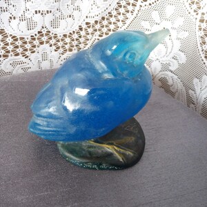 Pode incluir: Uma figurinha de p&aacute;ssaro de vidro azul com uma superf&iacute;cie texturizada detalhada. O p&aacute;ssaro est&aacute; empoleirado em uma base marrom texturizada.