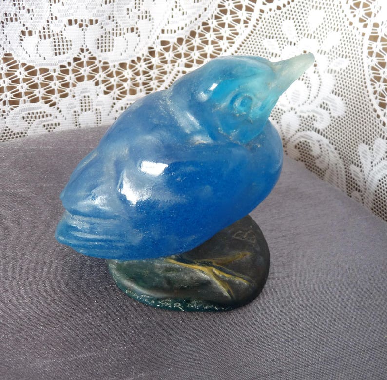 Pode incluir: Uma figurinha de p&aacute;ssaro de vidro azul com uma superf&iacute;cie texturizada detalhada. O p&aacute;ssaro est&aacute; empoleirado em uma base marrom texturizada.