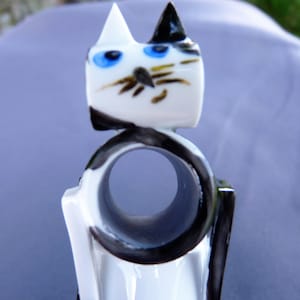 May include: A black and white ceramic cat figurine with blue eyes. The cat is sitting on a ring-shaped base with a hole in the center.
