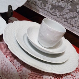 Puede incluir: Un juego de cuatro platos de porcelana blanca con un delicado diseño floral. Los platos están apilados uno encima del otro, con una taza de porcelana blanca sobre el plato más pequeño.