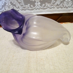 Puede incluir: Un jarrón de vidrio morado y blanco con una forma orgánica única. El jarrón tiene una abertura ancha y un cuello estrecho, y está hecho de un vidrio translúcido que permite que la luz brille a través de él.