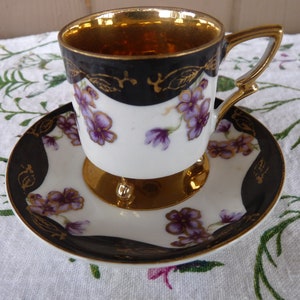 Puede incluir: Una taza de té y un platillo blancos con un borde negro y dorado y un diseño floral morado. La taza tiene un interior dorado y tres patas doradas.