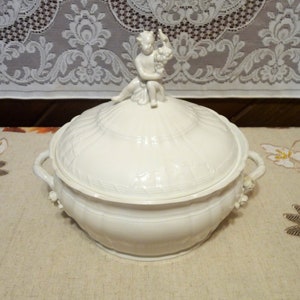 May include: White porcelain serving dish with a lid. The lid features a sculpted cherub figure holding grapes. The dish has two handles and intricate floral designs.