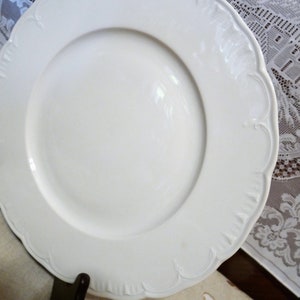 May include: White ceramic dinner plate with a scalloped edge and a raised rim. The plate has a delicate floral design.