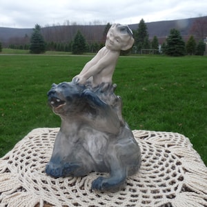 以下が含まれることがあります： いたずらっ子の小さな男の子が熊の背中に乗っている、磁器製の置物です。男の子は裸で、とがった耳をしています。熊は灰色で、青みがかった灰色の腹をしています。置物は白いレースのドイリーの上に置かれています。