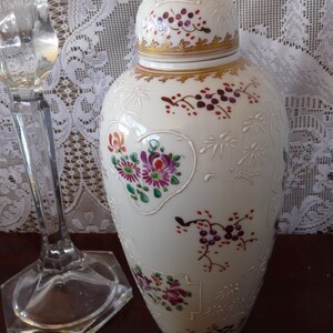 May include: A white porcelain vase with a lid, decorated with floral and vine patterns in shades of pink, purple, red, and green. The vase has a gold trim around the lid and base.