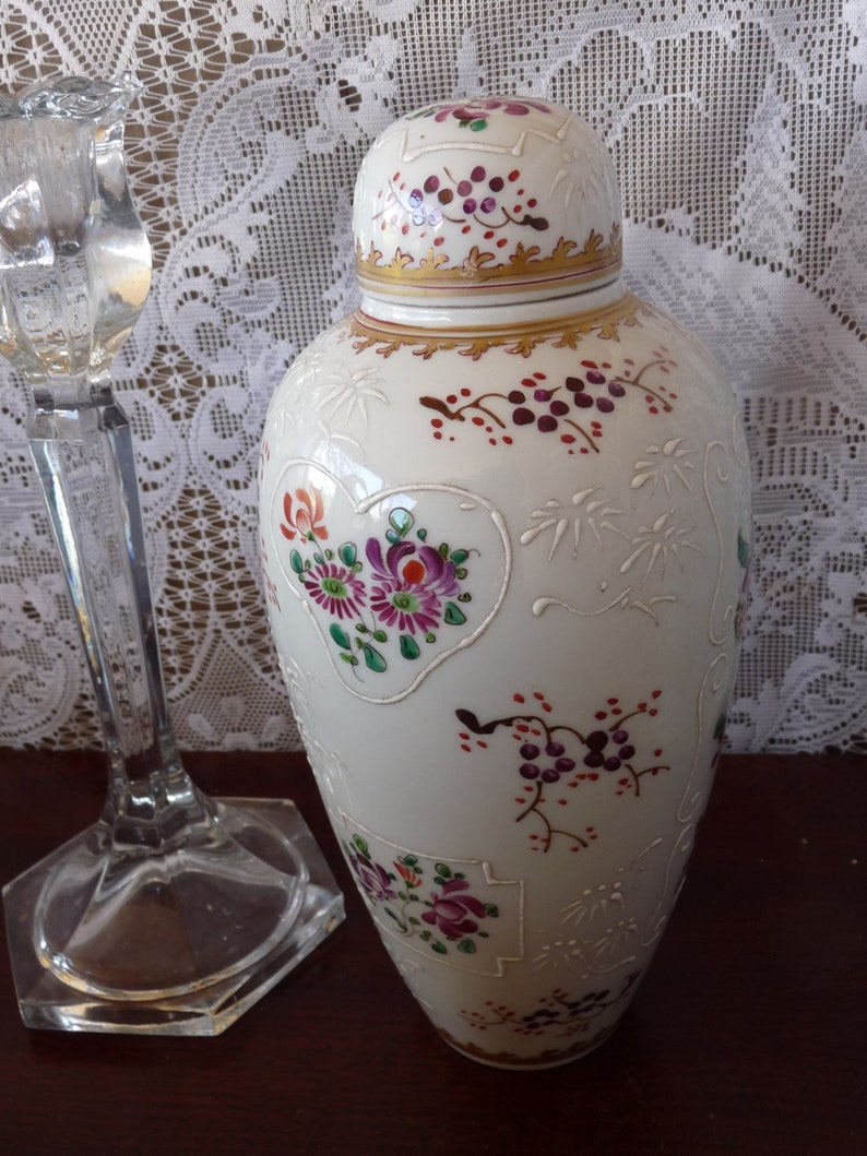 May include: A white porcelain vase with a lid, decorated with floral and vine patterns in shades of pink, purple, red, and green. The vase has a gold trim around the lid and base.