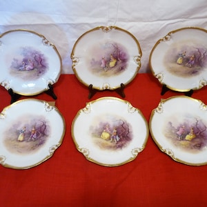 May include: Set of six white porcelain plates with gold trim and a painted scene of people in a garden.