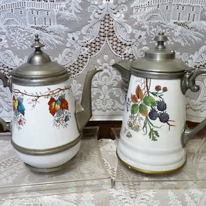 Puede incluir: Dos teteras antiguas de cerámica blanca con tapas y asas de metal. Una tetera presenta un diseño de vid con hojas coloridas, mientras que la otra tiene un patrón de moras y hojas. Ambas teteras se exhiben sobre soportes acrílicos transparentes.