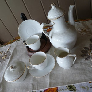 Könnte beinhalten: Ein weißes Porzellan-Tee-Set mit goldenen Verzierungen. Das Set enthält eine Teekanne, eine Milchkännchen, eine Zuckerdose und zwei Teetassen mit Untertassen.