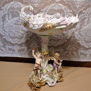 Puede incluir: Un centro de mesa de porcelana con un cuenco en forma de concha blanca y rosa, un diseño floral y dos figuras de querubines. Los querubines sostienen frutas y flores.