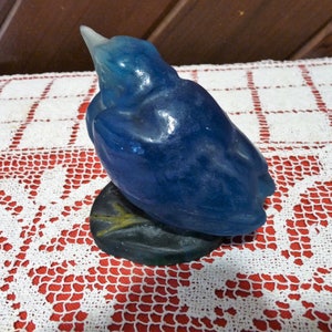 Pode incluir: Uma figurinha de vidro azul de um p&aacute;ssaro com um bico longo, empoleirado em uma base verde escura. O p&aacute;ssaro est&aacute; de costas para o observador, com a cabe&ccedil;a inclinada para tr&aacute;s.