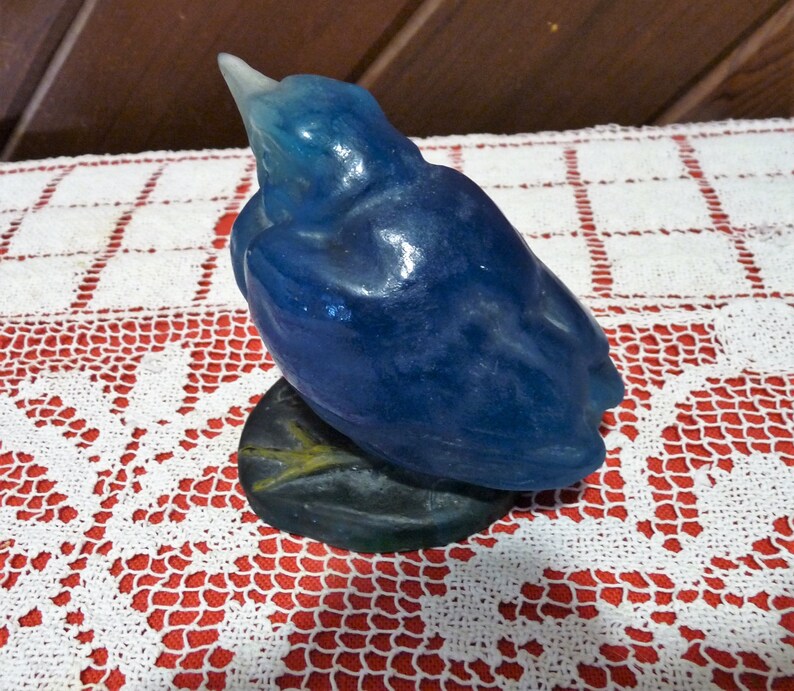 Pode incluir: Uma figurinha de vidro azul de um p&aacute;ssaro com um bico longo, empoleirado em uma base verde escura. O p&aacute;ssaro est&aacute; de costas para o observador, com a cabe&ccedil;a inclinada para tr&aacute;s.