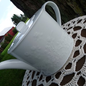 May include: White porcelain teapot with a floral design. The teapot has a handle and a spout. It is sitting on a crocheted doily.