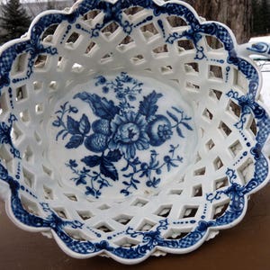 May include: A white porcelain basket with blue floral designs and a lattice pattern. The basket has two handles and is perfect for serving fruit or other small items.