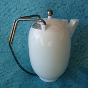 以下が含まれることがあります： 銀色の取っ手と蓋が付いた白い陶器製のティーポット。ティーポットの上部に銀色のバンドがあり、蓋には銀色のノブが付いています。