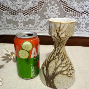 Puede incluir: Un jarrón de cerámica con un diseño de árbol en marrón y blanco. El jarrón tiene una base ancha y un cuello estrecho. Está sentado en una mesa con una lata de agua con gas de fresa y pepino.