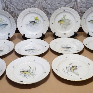 May include: A set of 12 white porcelain dinner plates with a gold rim. Each plate features a hand-painted design of a fish swimming in a pond with reeds and lily pads.