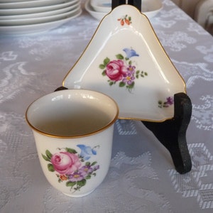Puede incluir: Un juego de taza y platillo de té de porcelana blanca con un diseño floral. La taza y el platillo están adornados con detalles dorados. El diseño floral presenta flores rosas, moradas y azules.