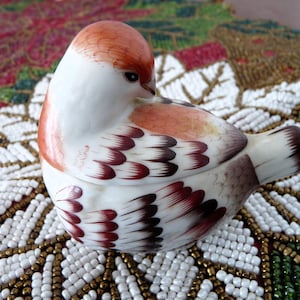 May include: A ceramic bird figurine with a white body, orange head, and brown and burgundy feather details. The bird is perched on a beaded surface with white, gold, and green beads. The figurine is a decorative item.