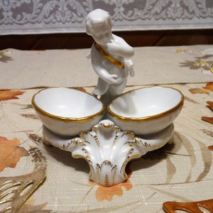 May include: White porcelain salt cellar with two bowls and a cherub figurine. The cherub is holding a golden ribbon and is standing on top of the salt cellar. The salt cellar has a gold trim around the edges of the bowls and the base.