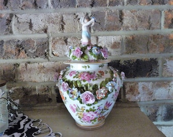 May include: A white porcelain vase with a floral design and a cherub figurine on top. The vase is decorated with pink, red, and yellow flowers.