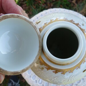 May include: A white porcelain jar with a lid, decorated with gold and red floral patterns. The jar has a wide opening and a smooth, white interior.