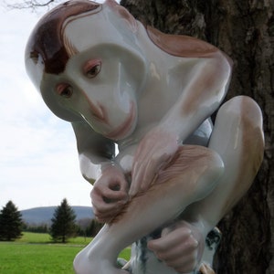 May include: A porcelain figurine of a monkey sitting on a tree stump. The monkey is painted in shades of brown, white, and pink. The monkey's eyes are closed and it is holding its foot with its hand.