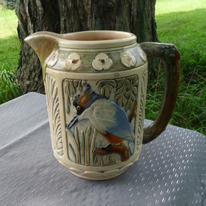 Puede incluir: Una jarra de cerámica con asa marrón y un diseño detallado de un pájaro en un entorno de bosque. La jarra está decorada con flores y hojas en tonos verdes, marrones y azules.