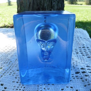 May include: A blue glass paperweight with a clear, sculpted human head inside. The head is facing forward and has a neutral expression.
