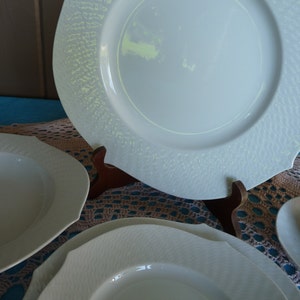 Puede incluir: Un conjunto de vajilla de porcelana blanca con un diseño de borde texturizado. El juego incluye platos, cuencos y una taza y platillo. Los platos tienen una forma única con bordes curvos. La vajilla se exhibe sobre una superficie azul.
