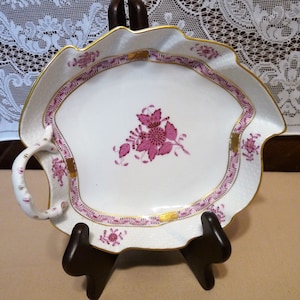 May include: A white porcelain leaf-shaped serving dish with a pink floral design and gold accents. The dish has a handle and is decorated with a pink floral pattern.