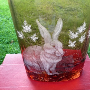 Puede incluir: Un jarrón de vidrio transparente con un tono marrón y naranja presenta un diseño grabado de un conejo blanco en un campo de flores blancas.