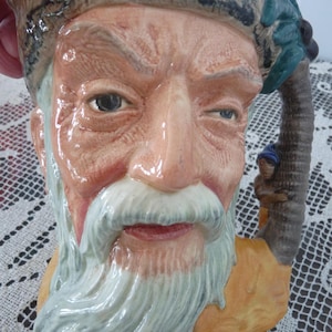 May include: A ceramic mug featuring a detailed, aged face with a long white beard and a fur hat. The handle is a palm tree with a small figure climbing it. The mug has a tan and brown color scheme.