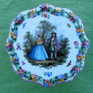 Puede incluir: Un plato decorativo de porcelana con borde festoneado. El centro presenta una escena pintada de dos figuras con vestimenta de época en un jardín. El borde está adornado con coloridos acentos florales y de hojas tridimensionales, con mariposas.