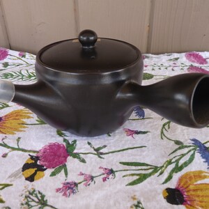 以下が含まれることがあります： 注ぎ口と蓋が付いた黒い陶器製のティーポット。ティーポットには白いシリコン製の注ぎ口カバーが付いています。
