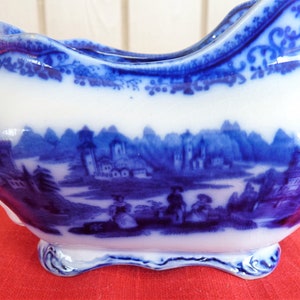 Puede incluir: Salsera de porcelana azul y blanca con asa y diseño escénico. La salsera tiene un borde festoneado y una base elevada.