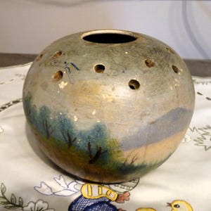 Pode incluir: Um vaso redondo de cerâmica com uma cena de paisagem pintada. O vaso tem um furo no topo e vários pequenos furos ao redor dos lados.