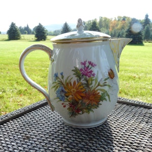 Puede incluir: Tetera de porcelana blanca con un diseño floral con flores rosas, amarillas y azules. La tetera tiene un borde dorado alrededor de la tapa y el pico.